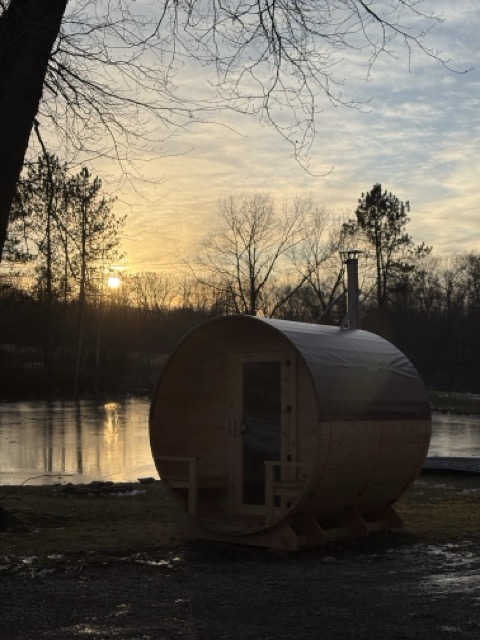 Cedar Ridge barrel sauna at sunset beside a frozen lake, bare winter trees and chimney pipe visible