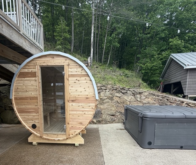 Cedar barrel sauna on a concrete patio alongside a hot tub, set against a wooded hillside with a deck above