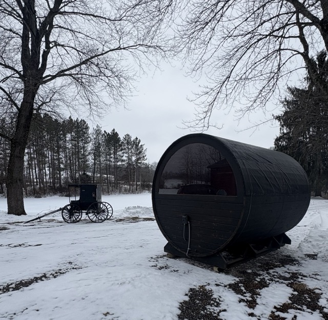 Dark-stained Cedar Ridge barrel sauna in a snowy yard with an Amish horse-drawn buggy in the background