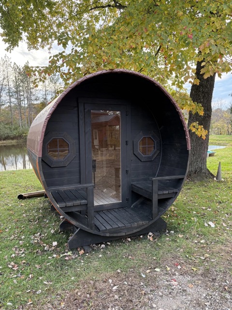 Dark-stained Cedar Ridge barrel sauna with entry porch and porthole windows beside a pond in autumn