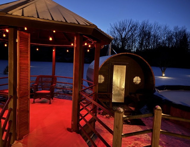 Cedar Ridge barrel sauna glowing at night beside a lit porch gazebo with string lights and wicker chair in the snow